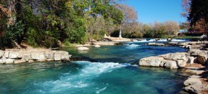 A tranquil river with clear, turquoise water flows over rocks and small cascades in San Marcos, surrounded by dense, green and autumn-colored trees under a bright blue sky. The serene setting includes some larger stones scattered along the riverbanks.