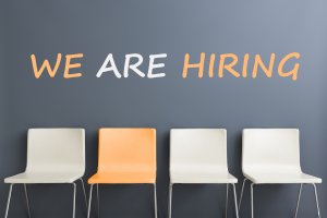 Four chairs are placed against a gray wall. The third chair from the left is orange, while the others are white. Above the chairs, the words "WE ARE HIRING" are written in large, bold letters, with "WE ARE" in white and "HIRING" in orange.