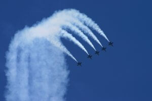 Five jet airplanes, reminiscent of the Blue Angels, fly in perfect formation with trails of smoke forming an arc against a clear blue sky. The planes are leaving distinct white vapor trails as they perform a synchronized aerial maneuver.