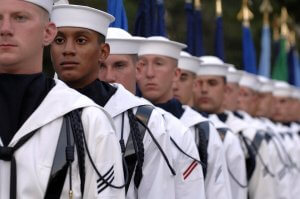 A group of Navy sailors, dressed in white uniforms and caps, stand in formation. The sailors are in focus, facing straight ahead, with various flags visible in the background. Their solemn and disciplined demeanor mirrors the precision of the Blue Angels.