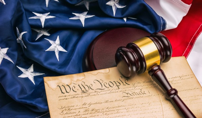 An American flag lays partially folded in the background with a wooden judge's gavel resting on top of it. In the foreground, a portion of the United States Constitution with the words "We the People" is visible, symbolizing leadership and justice intertwined.