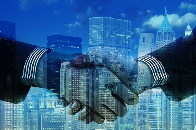 A double exposure image showing two hands shaking, symbolizing a business agreement and leadership, overlaid with skyscrapers and city buildings. The background features a modern cityscape with a predominantly blue hue.