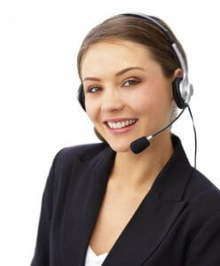 A smiling woman wearing a black blazer, a white top, and a headset with a microphone, stands against a plain white background. She appears to be a customer service representative or a call center agent.