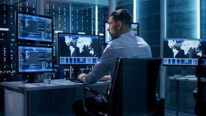 A man in a white shirt sits at a desk with multiple computer monitors displaying world maps and data, immersed in a cybersecurity environment. The room has a dark, modern aesthetic with a large server rack in the background, emphasizing its role in cyber defense.