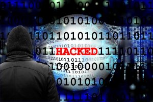 A person in a hooded jacket faces a digital globe with binary code in the background. The word "HACKED" is prominently displayed in red across the center of the image, symbolizing a cybersecurity breach or cyberattack.