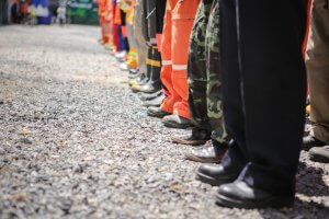 A row of people standing in line on gravel, photographed from the waist down. They are wearing various types of work pants and boots, including orange coveralls and camouflage pants, ready for an emergency situation.