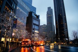 Cityscape at dusk featuring tall buildings with illuminated windows. Two emergency fire trucks with lights flashing are parked on a wet street lined with pedestrians and trees wrapped in twinkling lights. Traffic signals and urban infrastructure are visible.