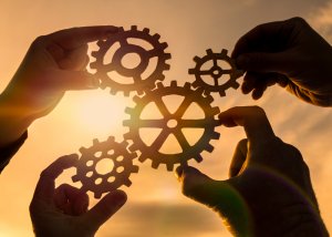 Five hands each hold a wooden cogwheel, fitting them together against a backdrop of a sunset, symbolizing teamwork and engagement. Warm sunlight flares through the gaps between the cogwheels, creating a golden glow.