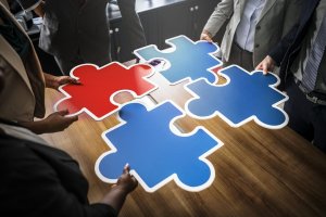 Four people holding large blue and red puzzle pieces in a boardroom setting, each piece forming part of a larger puzzle on a wooden table. The individuals are dressed in business attire, symbolizing collaboration and teamwork, fostering an environment of active engagement.