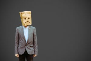 A person wearing a gray suit and blue shirt stands against a plain dark background. Their head is covered with a brown paper bag that has a sad face drawn on it, consisting of two black dots for eyes and a downturned curve for a mouth, as if ghosting the world around them.