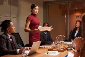 A woman in a red dress stands and speaks, holding papers, exuding leadership in the conference room. She is surrounded by colleagues seated at a table with laptops, notepads, and a glass pitcher. The atmosphere is professional and focused.