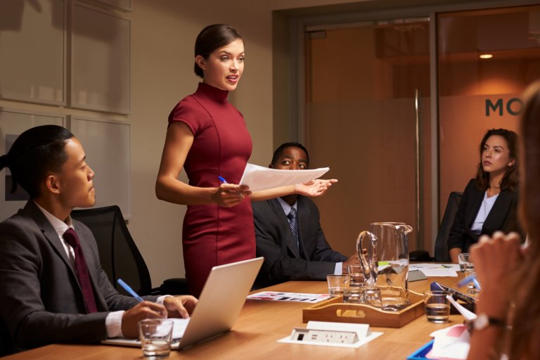 A woman in a red dress stands and speaks, holding papers, exuding leadership in the conference room. She is surrounded by colleagues seated at a table with laptops, notepads, and a glass pitcher. The atmosphere is professional and focused.
