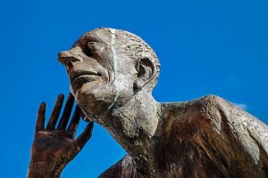 A bronze statue of a bald, older man with an elongated face and prominent nose, leaning forward and cupping his hand to his ear, as if trying to hear something. The statue, symbolizing attentive leadership, is set against a bright blue sky in the background.