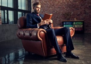A man dressed in a formal suit sits comfortably in a brown leather armchair, engrossed in reading a book. The setting, filled with potential, is an industrial-style room with brick walls, large windows, and a shelf with multicolored books in the background.