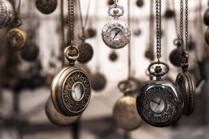 A collection of vintage pocket watches hangs by chains, showcasing intricate designs and metallic finishes in silver, bronze, and gold. The background is blurred, highlighting the potential of each detailed timepiece's craftsmanship in the foreground.
