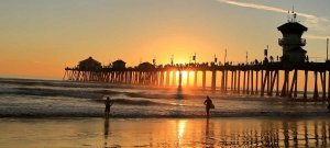 A sunset view of an Orange County beach with a pier extending into the ocean. People are walking on the pier, their silhouettes visible against the golden sky. Waves gently wash onto the shore where two individuals stand near the water's edge.