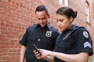 Two police officers in uniform stand in a brick alleyway. One of the police officers, holding a smartphone, appears focused on the screen. The other officer looks on with interest. Both officers have badges and patches on their uniforms.