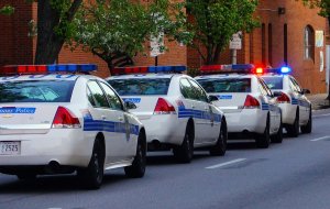 Four police cars with flashing lights are parked in a line on the side of the street. The vehicles, marked "Baltimore Police," stand ready as trees create a leafy backdrop and a brick building rises to the right. It appears to be daytime.
