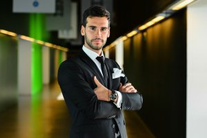 A confident man in a black suit and tie stands with his arms crossed, exuding an imposing presence. He has dark hair and a neatly trimmed beard, and he is looking directly at the camera. The background features a modern corridor with green and yellow walls, softly illuminated by ceiling lights.