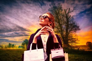 A stylish woman wearing dark sunglasses and a dark coat, exuding an air of presence, holds several shopping bags while looking up thoughtfully. The background features a dramatic sunset sky with clouds and silhouettes of trees.