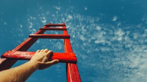 A person's hand holds onto a red ladder, climbing up against a backdrop of clear blue sky with scattered white clouds. The perspective highlights a sense of determination and the potential for reaching new heights.