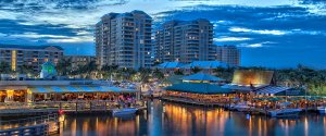A vibrant waterfront scene at dusk in Boynton with multi-story buildings in the background. The area is bustling with lively restaurants and shops along the water. Boats are docked along the waterfront, and the sky is painted with beautiful blue and orange hues.