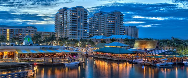 A vibrant waterfront scene at dusk in Boynton with multi-story buildings in the background. The area is bustling with lively restaurants and shops along the water. Boats are docked along the waterfront, and the sky is painted with beautiful blue and orange hues.