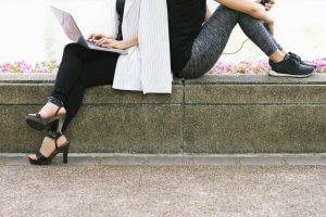 Two people sitting back-to-back on a stone bench, perfectly balanced in contrast. One wears black leggings, strappy heels, and uses a laptop, while the other sports grey leggings, sneakers, and holds a phone with earphones plugged in. Flowers and a path serve as their tranquil backdrop.