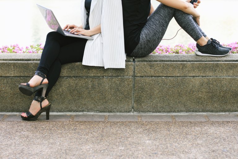 Two people sitting back-to-back on a stone bench, perfectly balanced in contrast. One wears black leggings, strappy heels, and uses a laptop, while the other sports grey leggings, sneakers, and holds a phone with earphones plugged in. Flowers and a path serve as their tranquil backdrop.