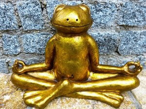A golden frog statue is seated in a cross-legged yoga pose with its hands resting on its knees in a meditative "okay" gesture, embodying serene balance. The statue is placed in front of a stone brick wall.