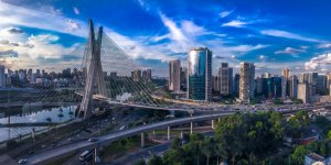 A panoramic view of a modern cityscape with a prominent cable-stayed bridge crossing a river, the marvel of any civil engineer. The skyline is filled with tall buildings and features a curving highway with bustling traffic. The sky is partly cloudy, enhancing the vibrant blue and green tones of the scene.