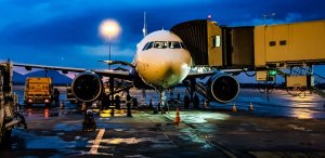 An airplane parked at an airport terminal gate during twilight, connected to a jet bridge designed by a civil engineer. The runway is wet and reflective after rainfall. The sky is dark and cloudy, with airport lights illuminating the area around the aircraft.