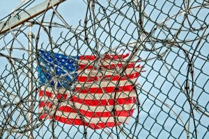 A U.S. flag is visible in the background behind a correctional facility's chain-link fence topped with barbed wire. The camera focuses on the fence, with the flag appearing slightly out of focus through the gaps in the wire. The sky is clear and blue.