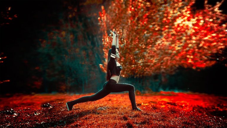 A person practices yoga outdoors in a lunge position with arms raised, embracing the tranquility of nature. They are immersed in a lush, vibrant environment with red and orange foliage, capturing the serene and colorful essence of autumnal culture.