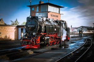A vintage black steam locomotive with red detailing sits idle on the track at a railway station with a brick building and control tower in the background. Steam is visible near the locomotive, and the platform is wet, reflecting the surroundings, evoking a nostalgic sense of railway culture.