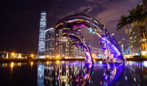 A vivid night cityscape features illuminated skyscrapers in the background. In the foreground, a striking sculpture of three silver dolphins appears to leap from a reflective pool, enhanced by colorful lighting. Palm trees and urban architecture frame the scene, showcasing a blend of culture and modernity.