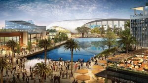 A lively outdoor scene in Inglewood features a crowd of people walking near a large reflective pool surrounded by trees, buildings, and patio seating. The background showcases modern architecture and a partially visible stadium, all under a sky with scattered clouds.