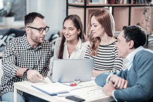 A group of four staff members engaged in a discussion around a table with a laptop and documents. One person is pointing at the screen, while the others listen attentively. The setting resembles a casual office or meeting space.