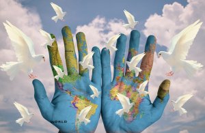 Two hands painted with a map of the world are held up against a cloudy sky. White doves are flying around the hands, symbolizing peace and unity, as if illustrating global projects envisioned in harmony.