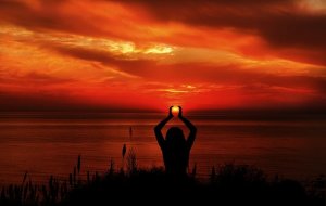 A silhouette of a person standing on a grassy ledge, projecting their hands upward to frame the setting sun against a vibrant, fiery orange and red sky over calm ocean waters.