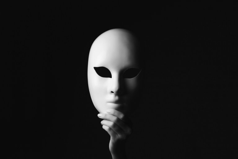 A white mask with black eye cutouts is held against a dark background by a hand positioned at the bottom, creating a mysterious and dramatic effect. The contrast between the mask and its dark surroundings highlights its features, embodying an enigmatic role within the scene.