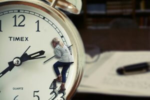 A person in casual attire appears to be climbing up the face of a large Timex clock, using the clock hands for support with impressive flexibility. The background is blurred, showing a desk with papers, books, and a pen. The image conveys a surreal representation of time and urgency.