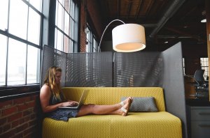A woman sits on a yellow couch, working on a laptop in a modern office space with brick walls and large windows. She has long blonde hair and is wearing a blue dress and white sandals. An overhead lamp brightens her flexible workspace, which is separated by a partition.