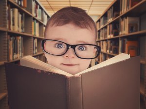 A young child with large black-rimmed glasses is peeking over the top of an open book, exhibiting a curious expression. In the background, a library staff member arranges rows of shelves filled with books, enhancing the scholarly atmosphere.