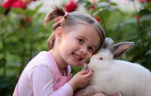 A young girl with blonde hair in pigtails, wearing a pink shirt, is smiling and holding a white and gray rabbit close to her face. She stands outdoors with green foliage and red flowers in the background, looking as if she could be part of the garden staff tending to the blossoms.