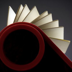 A visually striking image shows an open notebook with pages fanned out, captured from a unique angle. The foreground features a large, red circular shape resembling a rolodex that frames the bottom of the notebook, adding an abstract and artistic element to the composition.