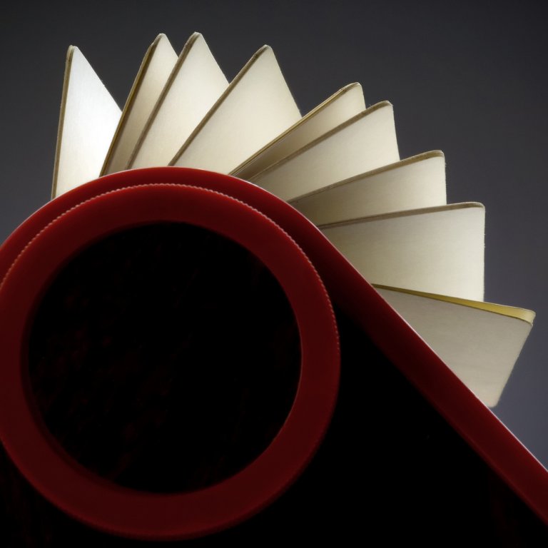 A visually striking image shows an open notebook with pages fanned out, captured from a unique angle. The foreground features a large, red circular shape resembling a rolodex that frames the bottom of the notebook, adding an abstract and artistic element to the composition.