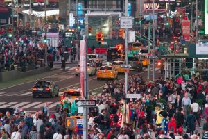 A bustling urban street scene at night with a dense crowd of pedestrians on sidewalks and crosswalks. Yellow taxis and other cars navigate through traffic as the passage of time feels palpable. Brightly lit signs, billboards, and storefronts illuminate the busy area.