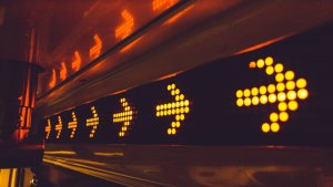 A series of bright yellow rightward-pointing arrows are displayed on an electronic sign against a dark background. The illuminated arrows serve as prominent directional indicators, resembling warning lights. The sign is placed indoors, encased in a metallic frame.