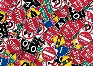 A chaotic pile of various road signs, including stop signs, do not enter signs, speed limit signs, wrong way signs, and directional signs interspersed with warning lights. The jumble creates a visually busy and colorful collage.